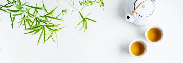 Asian tea concept, two white cups of tea and teapot surrounded with green tea dry leaves view from above, space for a text on white background.Long wide banner