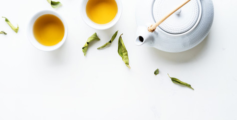 Asian tea concept, two white cups of tea and teapot surrounded with green tea dry leaves view from above, space for a text on white background. Long wide banner