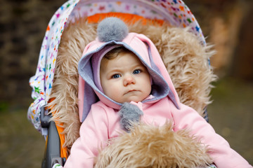 Canvas Print - Cute little beautiful baby girl sitting in the pram or stroller on autumn day. Happy smiling child in warm clothes, fashion stylish pink baby coat with bunny ears. Baby going on a walk with parents.