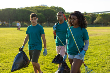 Mix raced group of volunteers carrying trash from city lawn. Young woman and men walking through city lawn, carrying rakes and plastic bags. Garbage removal concept