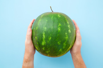 Young man hands holding one green watermelon on pastel blue table. Closeup.