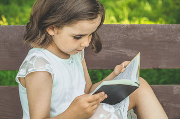 Wall Mural - Little Girl holds book in her hands. Reading the book in in outdoors. The girl on sitting on a bench, reading a book. Rest and reading