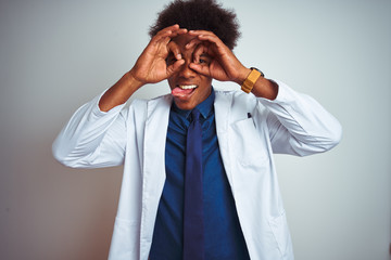 Poster - Young african american doctor man wearing coat standing over isolated white background doing ok gesture like binoculars sticking tongue out, eyes looking through fingers. Crazy expression.