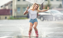 Cheerful Girl Jumping With White Umbrella In Dotted Red Galoshes. Hot Summer Day After The Rain Woman Jumping And Splashing In Puddle