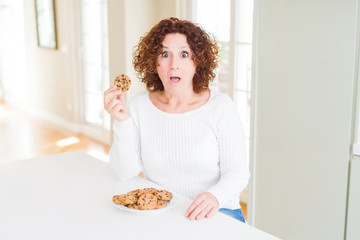 Poster - Senior woman eating chocolate chips cookies at home scared in shock with a surprise face, afraid and excited with fear expression