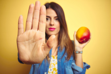 Wall Mural - Young beautiful woman eating fresh healthy mango over yellow background with open hand doing stop sign with serious and confident expression, defense gesture