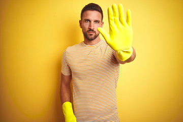 Wall Mural - Young handsome man wearing cleaning gloves for housework over isolated yellow background with open hand doing stop sign with serious and confident expression, defense gesture