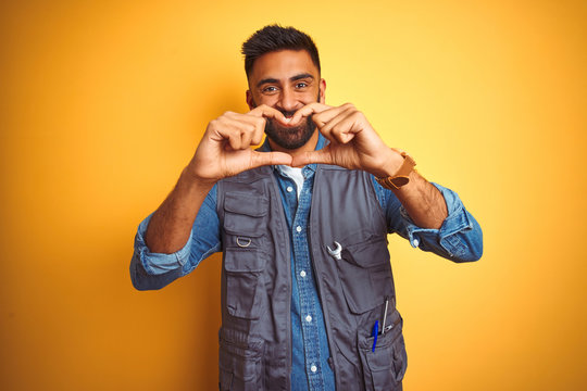 Handsome indian technician man wearing uniform over isolated yellow background smiling in love showing heart symbol and shape with hands. Romantic concept.