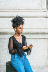 Wall Mural - Young African American Woman with afro hairstyle, wearing mesh sheer long sleeve shirt blouse, blue jeans, sitting on street in New York City, listening music with earphone and cell phone, texting..