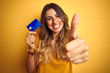 © Krakenimages.com - Young beautiful woman holding credit card over yellow isolated background happy with big smile doing ok sign, thumb up with fingers, excellent sign
