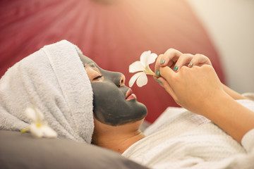 Young Asian woman beautiful in clay mask she is smell a flower. Woman spa face mask in spa uniform.