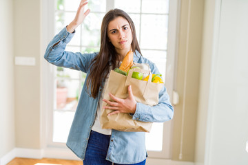 Poster - Beautiful young woman holding paper bag full of healthy groceries annoyed and frustrated shouting with anger, crazy and yelling with raised hand, anger concept