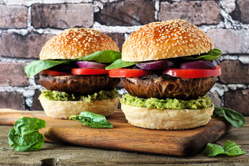 Wall Mural - Portobello mushroom vegan burgers with avocado, tomato and spinach and onion on a wood serving board against a dark brick background