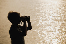 Closeup View Of Black Silhouette Of Little Kid Standing Outdoor At Blurry Golden River Water Background. Boy Looking In Old Vintage Binocular. Horizontal Color Photography.
