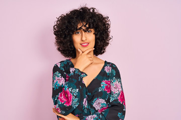 Young arab woman with curly hair wearing floral dress over isolated pink background looking confident at the camera with smile with crossed arms and hand raised on chin. Thinking positive.