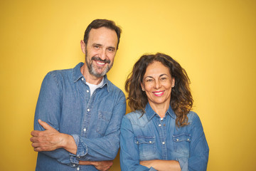 Beautiful middle age couple together standing over isolated yellow background happy face smiling with crossed arms looking at the camera. Positive person.