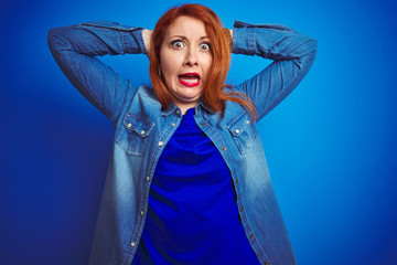 Wall Mural - Young beautiful redhead woman wearing denim shirt standing over blue isolated background Crazy and scared with hands on head, afraid and surprised of shock with open mouth