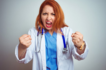 Wall Mural - Young redhead doctor woman using stethoscope over white isolated background angry and mad raising fists frustrated and furious while shouting with anger. Rage and aggressive concept.