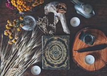 Flat Lay Of Witch's Altar Space With A Grimoire (vintage Book From 1837) And Other Various Items - Dried Flowers Nature Elements, Candles, Ink Bottle With Black Feather Next To It On Dark Wooden Table