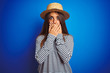 Young beautiful woman wearing navy striped t-shirt and hat over isolated blue background shocked covering mouth with hands for mistake. Secret concept.
