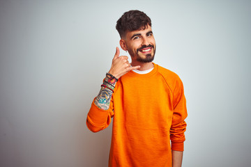 Young man with tattoo wearing orange sweater standing over isolated white background smiling doing phone gesture with hand and fingers like talking on the telephone. Communicating concepts.