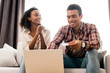 © LIGHTFIELD STUDIOS - handsome man and african american woman sitting on sofa while husband looking at laptop and holding credit card and wife looking at man with clenched hands