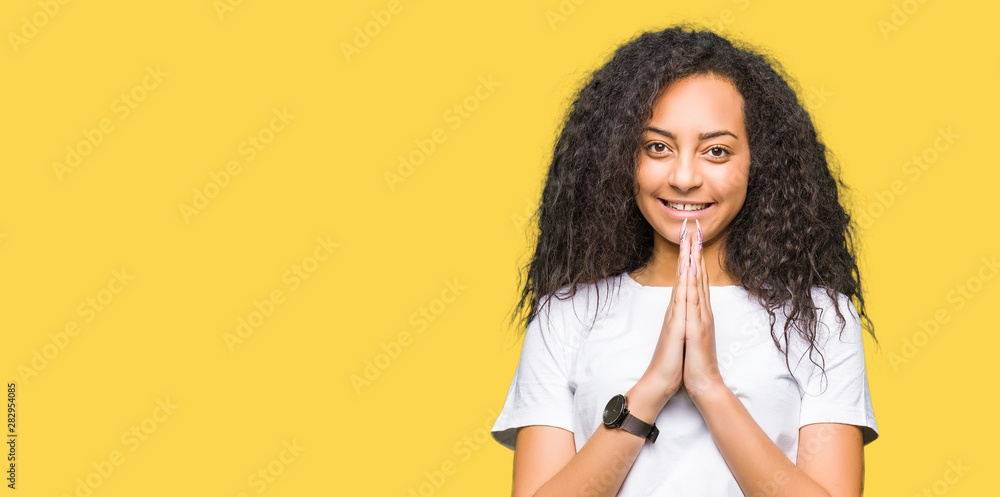 Young beautiful girl with curly hair wearing casual white t-shirt praying with hands together asking for forgiveness smiling confident.