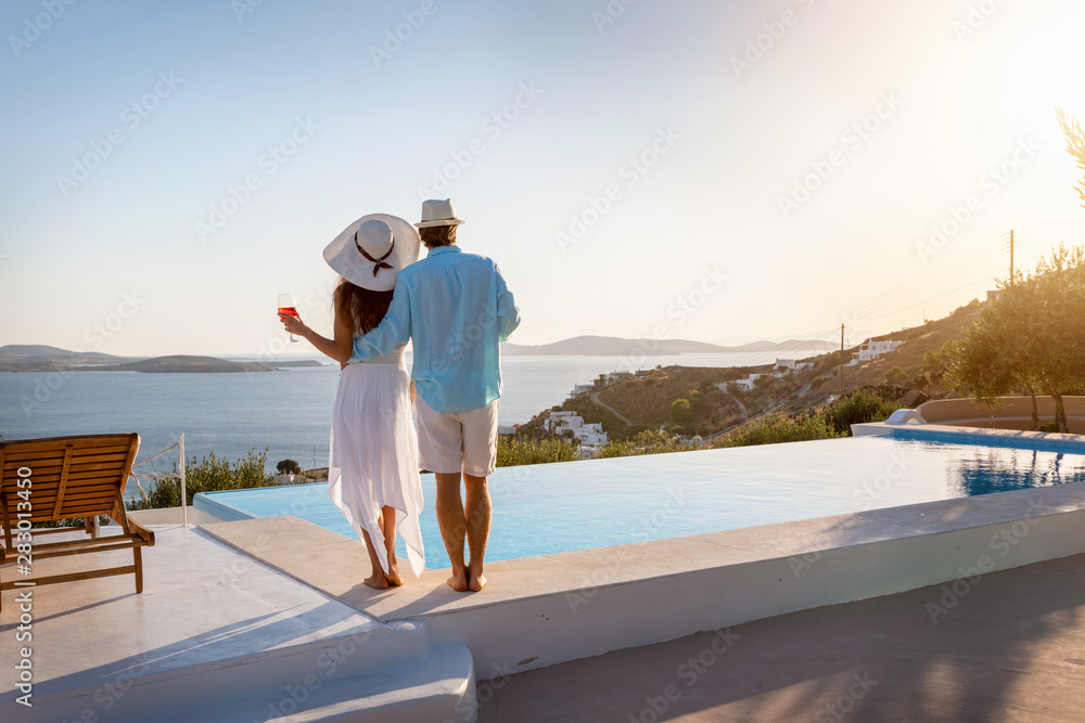 Romantisches Paar steht am Pool und genießt den Sonnenuntergang bei einem Glas Aperitif während des Sommerurlaubes - obrazy, fototapety, plakaty 
