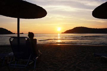 Happy woman have fun under umbrella on sandy beach  in Greece at sunset. Silhouette of woman sit under parasol and watch sunset