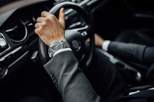 Close Up Top View Of  Man's Watch In Black Suit Keeping Hand On The Steering Wheel While Driving A Luxury Car.