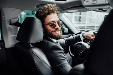 Wall Mural - Handsome young businessman in full suit smiling and driving a new car. View from behind.