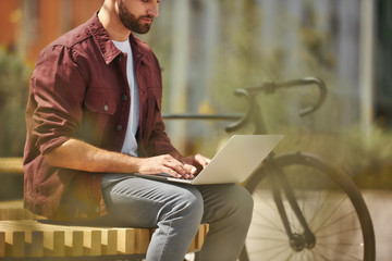 Wall Mural - I am always in touch. Side view of young man with stubble in casual clothes working on laptop while sitting on the bench near his bicycle