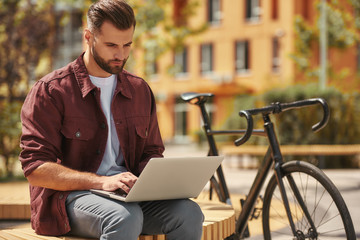 Wall Mural - Modern blogger. Portrait of young man with stubble in casual clothes working on laptop while sitting on the bench near his bicycle