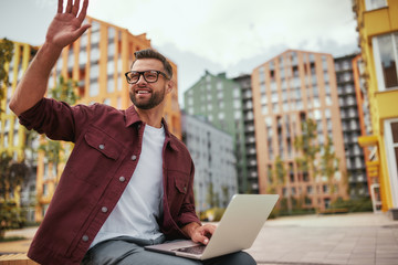 Wall Mural - I am here Young and cheerful handsome man with stubble in casual clothes and eyeglasses is waving to a friend while sitting on the bench with laptop outdoors