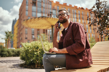 Wall Mural - Talking with client. Young handsome man with stubble in casual clothes talking by mobile phone while working on laptop on the bench outdoors