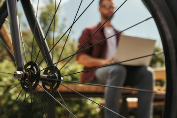 Wall Mural - Closeup of a bicycle wheel and man behind it working on laptop while sitting on the bench outdoors