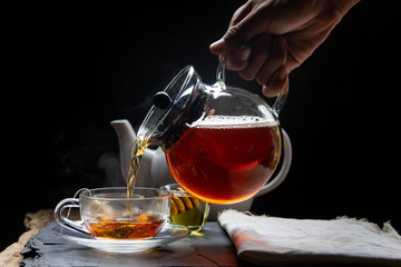 Hands are catching hot tea in the teapot and teacup with steam on reviving the wood and black background.