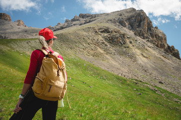 Wall Mural - View from the back Girl athlete in a red cap sunglasses and a yellow backpack stands on a green slope against the background of the epic cliffs of the Caucasus