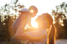 Backlight Silhouette Of A Mother Raising Her Kid Son At Sunset With The Sun In The Middle. Happy Mom Holding Her Cute Baby On Sunset Background
