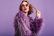 © Look! - Elegant pale girl wih bright pink lips plays with short curly hair. Romantic woman in fluffy purple jacket posing in studio in sunglasses.