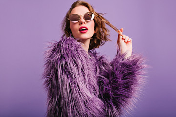 Elegant pale girl wih bright pink lips plays with short curly hair. Romantic woman in fluffy purple jacket posing in studio in sunglasses.