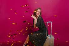 Pleased Stylish Lady Dreamy Posing On Purple Background Before Upcoming Trip. Elegant Female Traveler Sitting On Suitcase Under Golden Confetti.