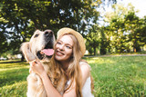 Fototapeta Zwierzęta - beautiful girl in white dress and straw hat hugging golden retriever while sitting on meadow, smiling and looking at dog