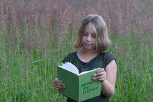  Little Girl Reads A Book Outside, Stands In A Green Dress In The Summer In The Field And Holds A Book In Her Hands