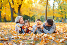 Happy Family Lying On Fallen Leaves, Playing And Having Fun In Autumn City Park. Children And Parents Together Having A Nice Day. Bright Sunlight And Yellow Leaves On Trees, Fall Season.