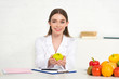 © LIGHTFIELD STUDIOS - front view of smiling dietitian in white coat holding apple at workplace
