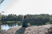 A Young Woman Sits On The Edge Of A Cliff Above A Quarry Filled With Water, Holding A Camera In Her Hands On A Summer Day And Takes Pictures Of The Landscape. Traveling As Lifestyle