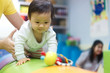 © Kawee - Young little Asian cute baby crawling on circular cushion practicing balance the body teaching by her preschool teacher in gym. The kid looking at toy and trying to catch it. Play and learn concept.