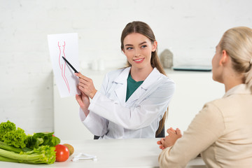 Wall Mural - smiling dietitian in white coat showing paper with perfect body image to patient