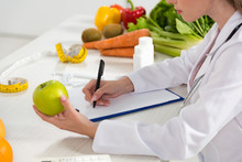 Cropped View Of Dietitian In White Coat Holding Green Apple And Writing In Clipboard